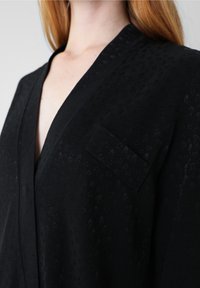 Gros plan d'une personne portant un blazer texturé noir avec une petite poche poitrine et un col en V profond.