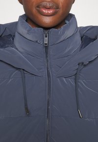 Navy puffer jacket featuring a high collar, front zipper, and adjustable drawstrings. Smooth texture with quilted detailing on the body.