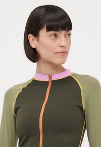 Wetsuit a maniche lunghe in verde oliva con accenti verde chiaro e lavanda, dotato di una zip frontale e dettagli di cucitura in contrasto.