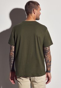 Camiseta de manga corta en verde oliva con cuello redondo, hecha de una tela suave, que presenta un ajuste relajado y un dobladillo limpio. Tatuajes visibles en los brazos.