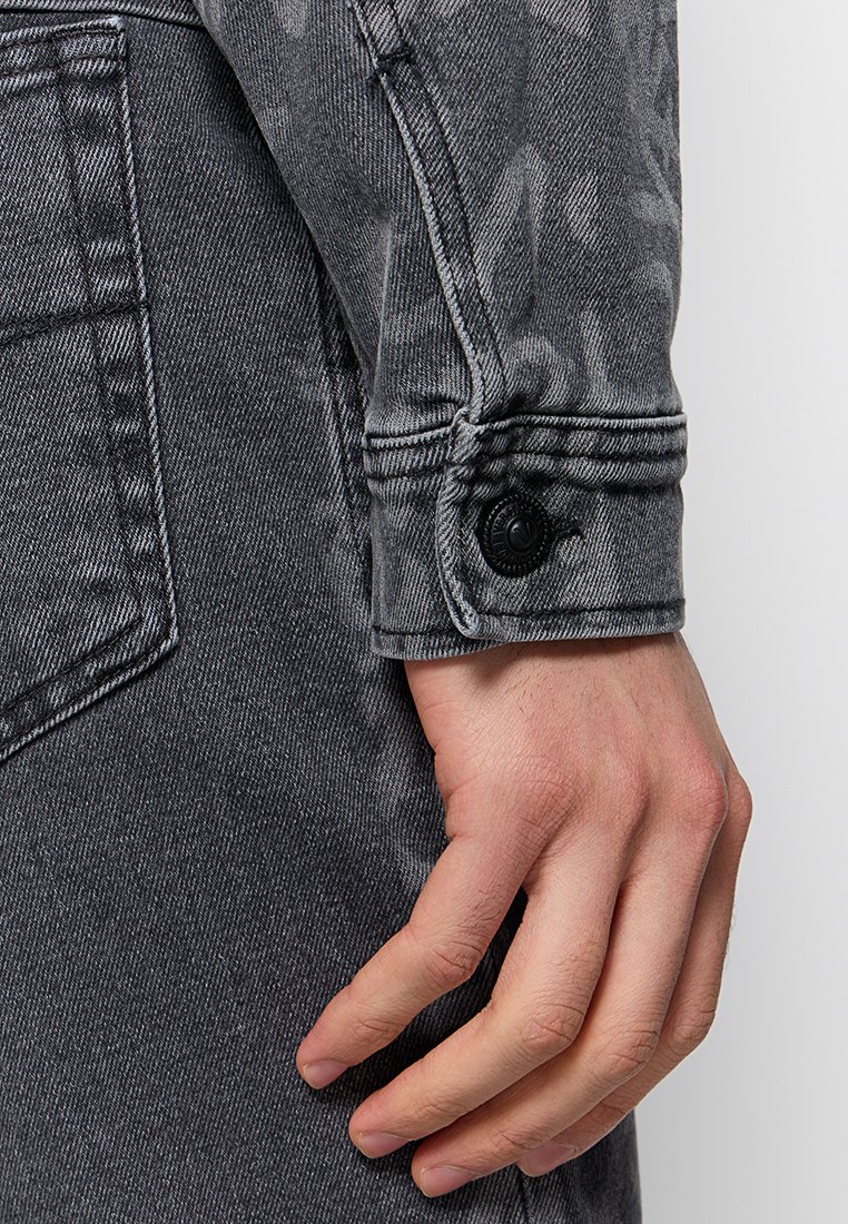 Saz para la manga de una chaqueta de mezclilla negra con un patrón texturizado, cierre de botón y costuras visibles. Se muestra una vista cercana del puño.