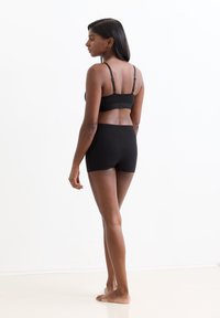 Frau steht barfuß seitlich, um ihren Rücken zu zeigen, trägt einen schwarzen verstellbaren BH und schwarze hochtaillierte Shorts vor einem weißen Hintergrund.