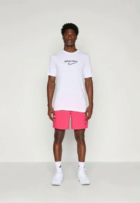 Balts Nike Pro T-krekls ar melnu logo, apvienots ar rozā šortiem ar logo, un baltiem Nike sporta apaviem. Vīrietis stāv neitrālā pozā.