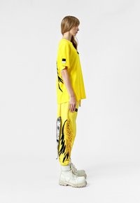 Camiseta amarilla de gran tamaño con un diseño gráfico negro en la parte trasera, combinada con pantalones a juego que presentan acentos texturizados y botas blancas.