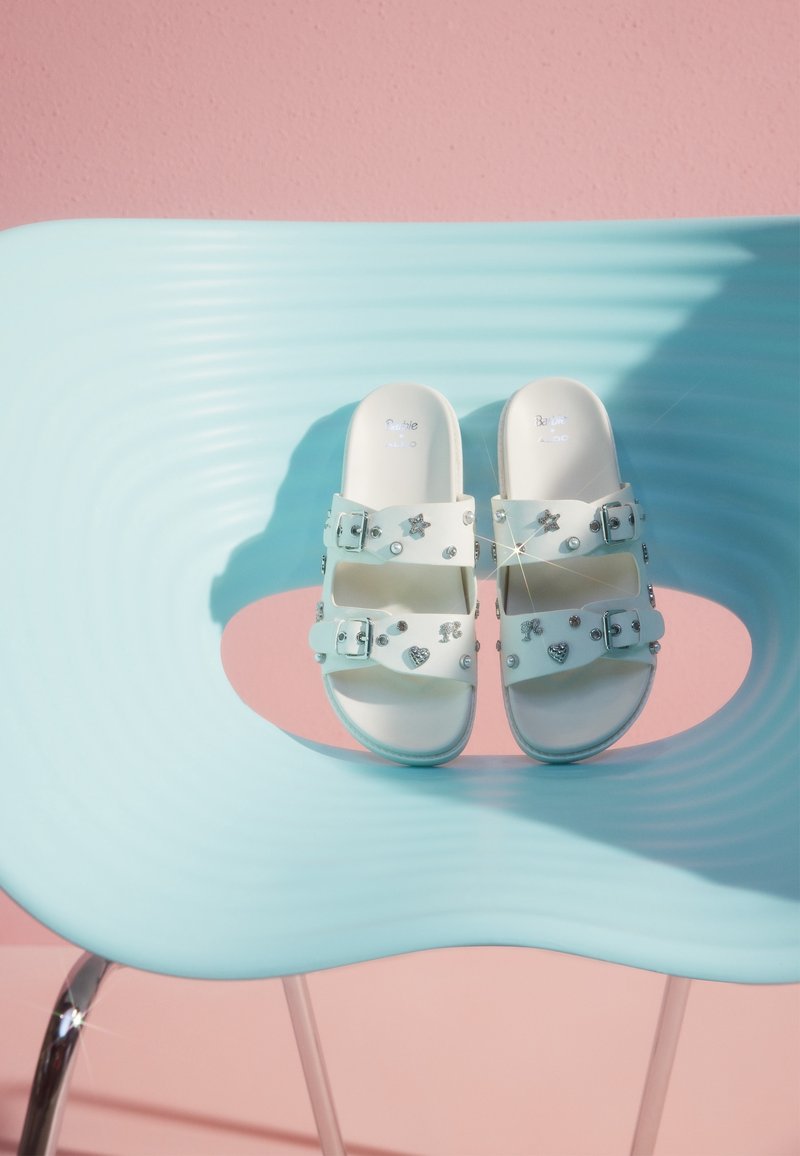 Sandales blanches à glissière avec double bride à boucle, ornées d'accents argentés et de divers motifs de cœurs et d'étoiles, posées sur une chaise turquoise texturée.