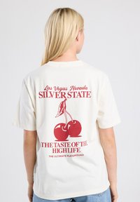 Cream-colored t-shirt with short sleeves, featuring red graphic text and cherry design on the back, smooth cotton texture.
