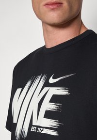 Camiseta de algodón negra con un gran logo blanco de Nike con acentos de pincel. Cuello redondeado y mangas cortas. Textura suave.