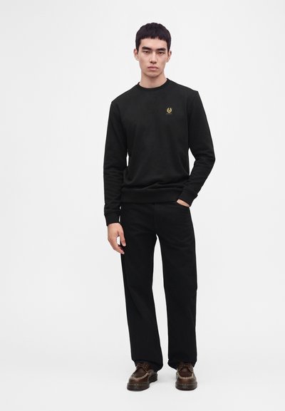 Jeune homme debout avec une main dans la poche, portant un sweat-shirt noir, un pantalon noir et des chaussures marron sur un fond blanc uni.