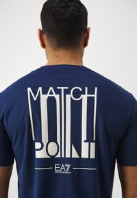 Tamsiai mėlyna marškinėliai su ryškiu baltu grafiniu tekstu "MATCH POINT" ir "EA7" logotipu nugaroje, pagaminti iš lygios, kvėpuojančios medžiagos.