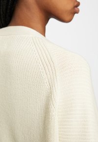 Sweter kardigan z długim rękawem w kolorze kremowym, charakteryzujący się teksturowanym materiałem, prążkowanymi detalami, szerokim kołnierzem oraz luźnym krojem.