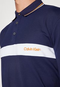 Ναυτικό πόλο μπλουζάκι με λευκή οριζόντια ρίγα και πορτοκαλί λογότυπο "Calvin Klein". Διαθέτει γιακά και τρία κουμπιά.