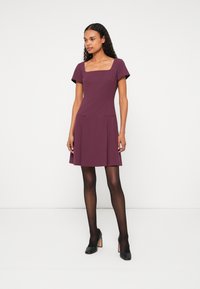 Rochie burgundy, cu croi prins pe talie și fustă evazată, având decolteu pătrat, mâneci scurte, buzunare laterale și un skirt plisat, purtată cu tocuri negre.