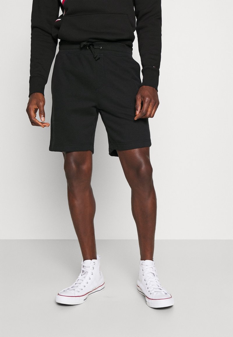 Mannequin masculin portant un short noir à cordon, un sweat-shirt noir et des baskets blanches montantes, debout sur un sol gris devant un mur blanc.