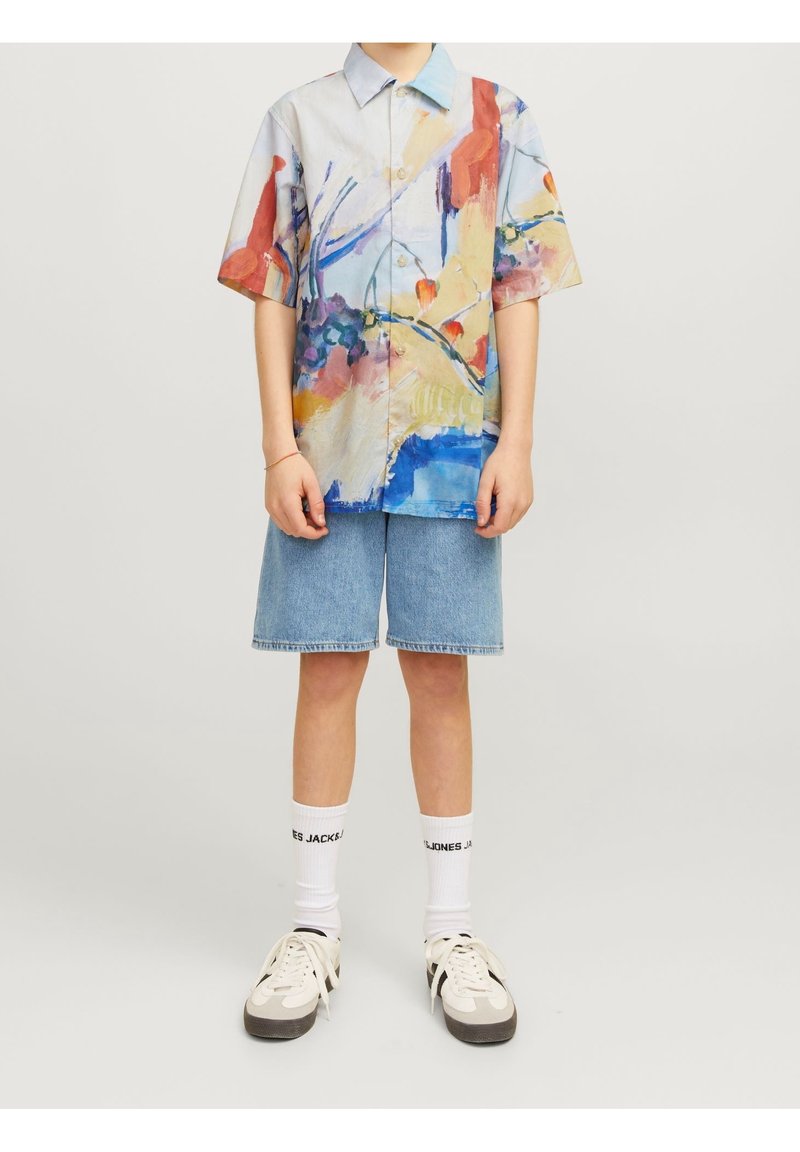 Henkilö, joka on pukeutunut värikkääseen abstraktiin lyhythihaiseen paitaan, vaaleansinisiin farkkushortseihin, valkoisiin nilkkasukkiin, joissa on mustaa tekstiä, ja valkoisiin tenkkuihin.