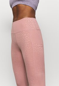 Růžové legíny s leopardím potiskem, které mají vysoký pas, strukturovanou tkaninu a boční kapsu. Design přechází z jednobarevného na vzorovaný.