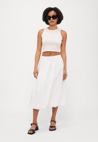 Lagani, bez rukava bež crop top, u kombinaciji s bijelom, lepršavom midi suknjom s elastičnom trakom. Crne kvadratne sunčane naočale i sandale.