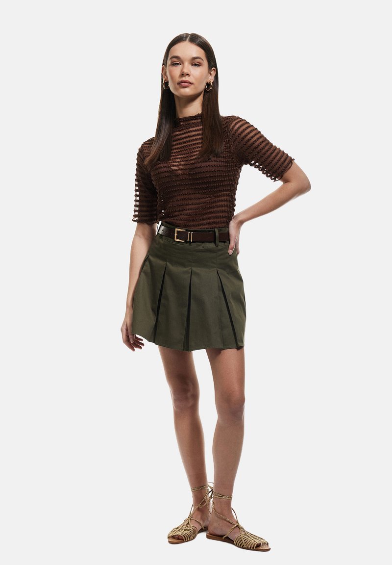 Top de punto marrón texturizado con mangas cortas, combinado con una falda plisada verde. Un cinturón marrón acentúa la cintura. Calzado: sandalias con tiras.