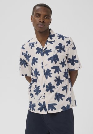 Ung mand med kort hår iført en kortærmet skjorte med store blå blomsterprint og mørke shorts, stående med hænderne på ryggen.