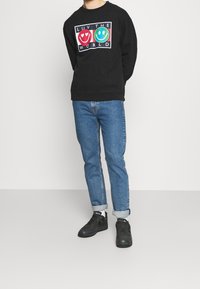 Felpa nera con una grafica colorata che recita "LUV THE WORLD" con faccine sorridenti, abbinata a jeans blu e scarpe da ginnastica nere.