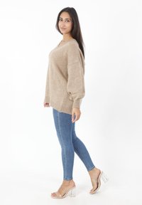 Beige oversized gebreide trui met lange mouwen, gecombineerd met strakke blauwe jeans en doorzichtige hakken, staand tegen een witte achtergrond.