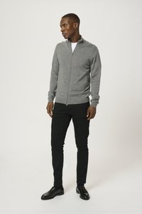 Grå zip-up-tröja i mjukt material, långa ärmar med ribbade manschetter, bärs över en vit skjorta, ihop med svarta byxor och skor.