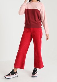 Karmesinrote Corduroy-Hosen mit weitem Bein kombiniert mit einem sweatshirt in rot und rosa mit Farbabstufungen. Schwarze und rosa Sportschuhe runden das Outfit ab.