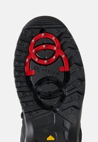 Černá gumová podešev s výrazným vzorkem běhounu. Obsahuje červené a černé kruhové detaily a zabudované kovové hroty pro lepší přilnavost.