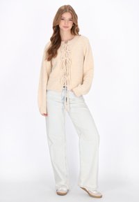 Beiger Strickpullover mit Schnürdetail auf der Vorderseite, langen Ärmeln und weichem Griff, kombiniert mit hellblauen weiten Jeans und hellen Schuhen.