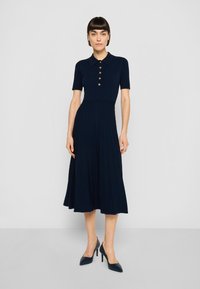 Tumši zila ribota trikotāžas kleita ar īsām piedurknēm, apkakli un zelta pogu akcentiem. Svārki ar vertikālām locītēm uz svārkiem. Valkāta kopā ar augstpapēžu kurpēm ar noapaļotiem purngaliem.