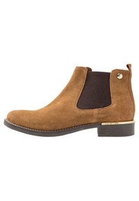 Stivale Chelsea in suede marrone con pannello laterale elasticizzato scuro, tacco basso impilato e piccolo dettaglio dorato vicino al tacco e alla linguetta posteriore.