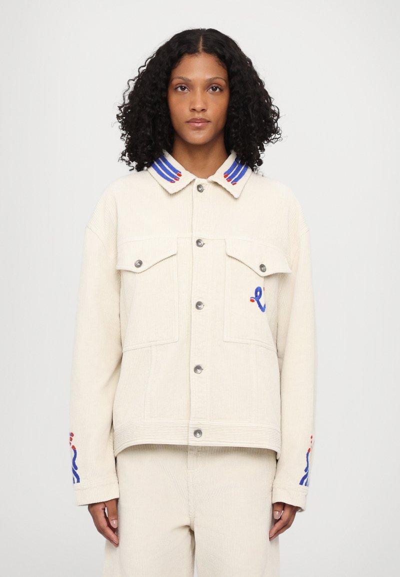 Beige fløjlsk jacket med knaplukning, to brystlommer og en farverig stribet krave. Har blå og røde broderede detaljer.