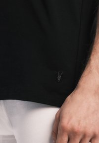 Black cotton t-shirt with a round neckline. Features a subtle, small embroidered design at the hem. Smooth texture and relaxed fit.