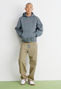 Grå hoodie med en frontlomme og et sort logo, parret med lysebeige bukser og hvide sneakers med røde accenter.