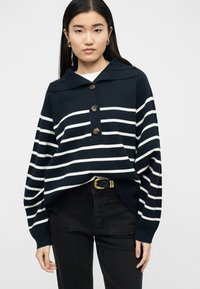 Jeune femme aux longs cheveux bruns portant un pull boutonné à rayures bleu marine et blanches, un jean noir et une ceinture noire avec une boucle dorée.