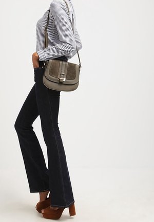 Sac bandoulière à rabat en cuir vert olive discret avec une texture subtile, ferrures argentées et sangle ajustable, accompagné d'un jean évasé foncé.