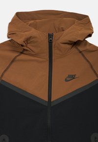 Brūns un melns Nike jaka ar kapuci, ar rāvējslēdzēju un slīpu krāsu bloku dizainu. Ietver logotipu un teksturētu neilona audumu.