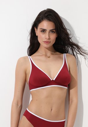 Donna con i capelli lunghi e scuri che indossa un bikini rosso con bordi bianchi, in piedi davanti a uno sfondo bianco, che guarda fiduciosa verso la fotocamera.