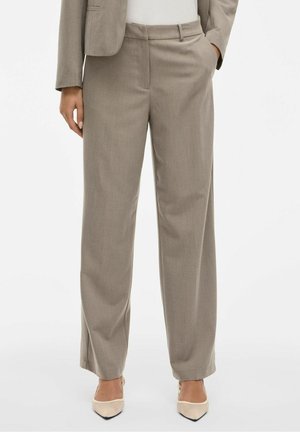 Vrouw draagt beige broek met wijde pijpen en bijpassend blazer, met wit topje en beige puntige schoenen, staand met één hand in de zak.