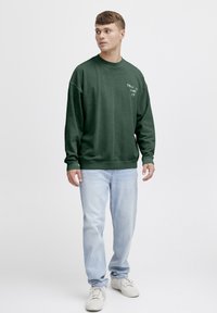 Sudadera verde oscura con cuello redondo, con texto blanco "DILO COMO ES" en el pecho. Combinada con jeans azul claro y zapatillas blancas.
