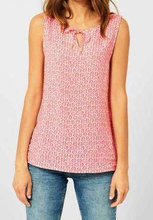 Frau trägt ärmelloses rosa gemustertes Oberteil mit Schlüssellochausschnitt und blaue Jeans, steht vor einem schlichten hellen Hintergrund.