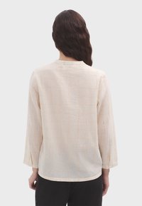 Beige bluse mit Gittermuster aus leichtem Stoff. Mit Rundhalsausschnitt, langen Ärmeln und einem lockeren Schnitt mit seitlichen Schlitzen.