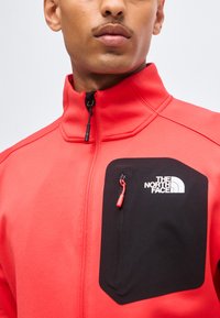 Homme portant une veste rouge zippée avec une poche poitrine zippée noire avec le logo The North Face en blanc.