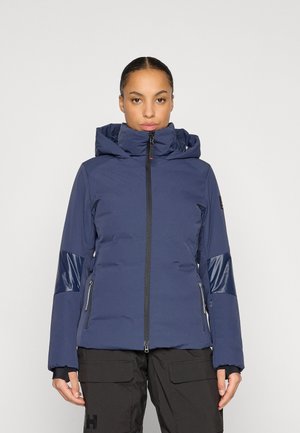 Woman wearing a blue hooded winter jacket with black zipper and pockets, paired with black cargo pants against a plain background.