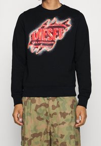 Sudadera negra con un logo gráfico en rojo y blanco en el pecho, que presenta un cuello y puños acanalados. Combinada con pantalones cortos de camuflaje.