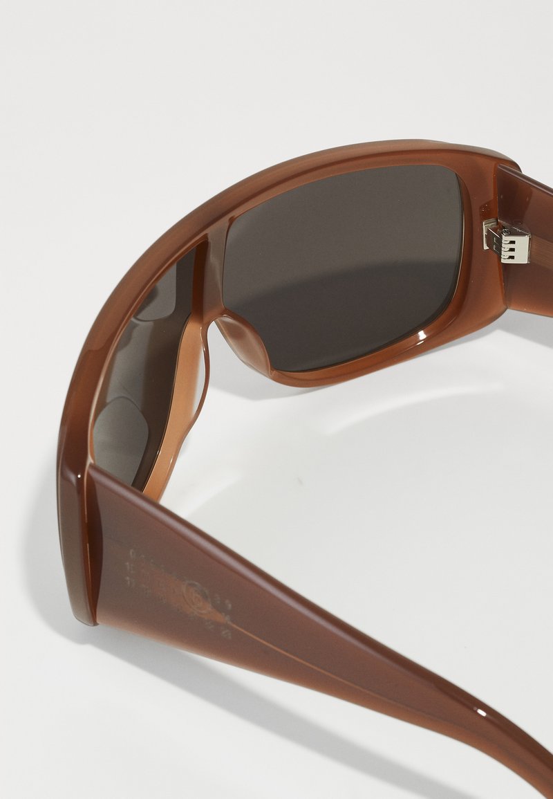 Lunettes de soleil en plastique brun avec des verres foncés, une finition lisse et un design enveloppant. Comprend un détail de charnière en métal sur la branche.