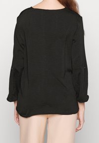 Kvinna iklädd en lös svart långärmad tröja och ljusbeiga byxor, sedd från ryggen mot en enkel bakgrund.