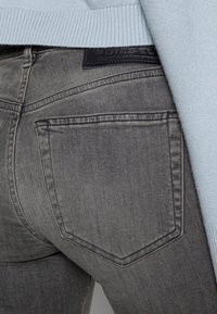 Jeans de mezclilla gris con un acabado texturizado. Presenta una forma distintiva en el bolsillo trasero y un parche con el logo negro cerca de la cinturilla. Suéter azul claro visible.