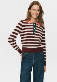Lächelnde Frau mit langen Haaren, die einen gestreiften Pullover mit bordeauxroten und weißen Streifen, einem korallenfarbenen Kragen und einer dunkelblaue Jeans trägt, steht vor einem weißen Hintergrund.