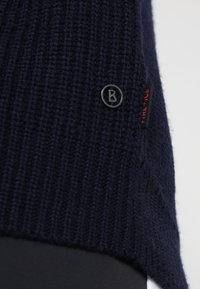 Pull en maille bleu marine avec texture côtelée, arborant un logo circulaire noir et une petite étiquette rouge ; coupe décontractée avec une fente sur le côté.