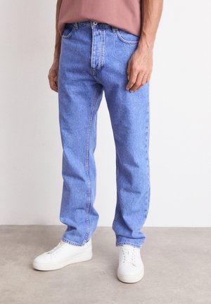 Mann trägt hellblaue, gerade geschnittene Jeans, weiße Turnschuhe und ein gedecktes rosa Hemd vor einem einfarbigen Hintergrund.